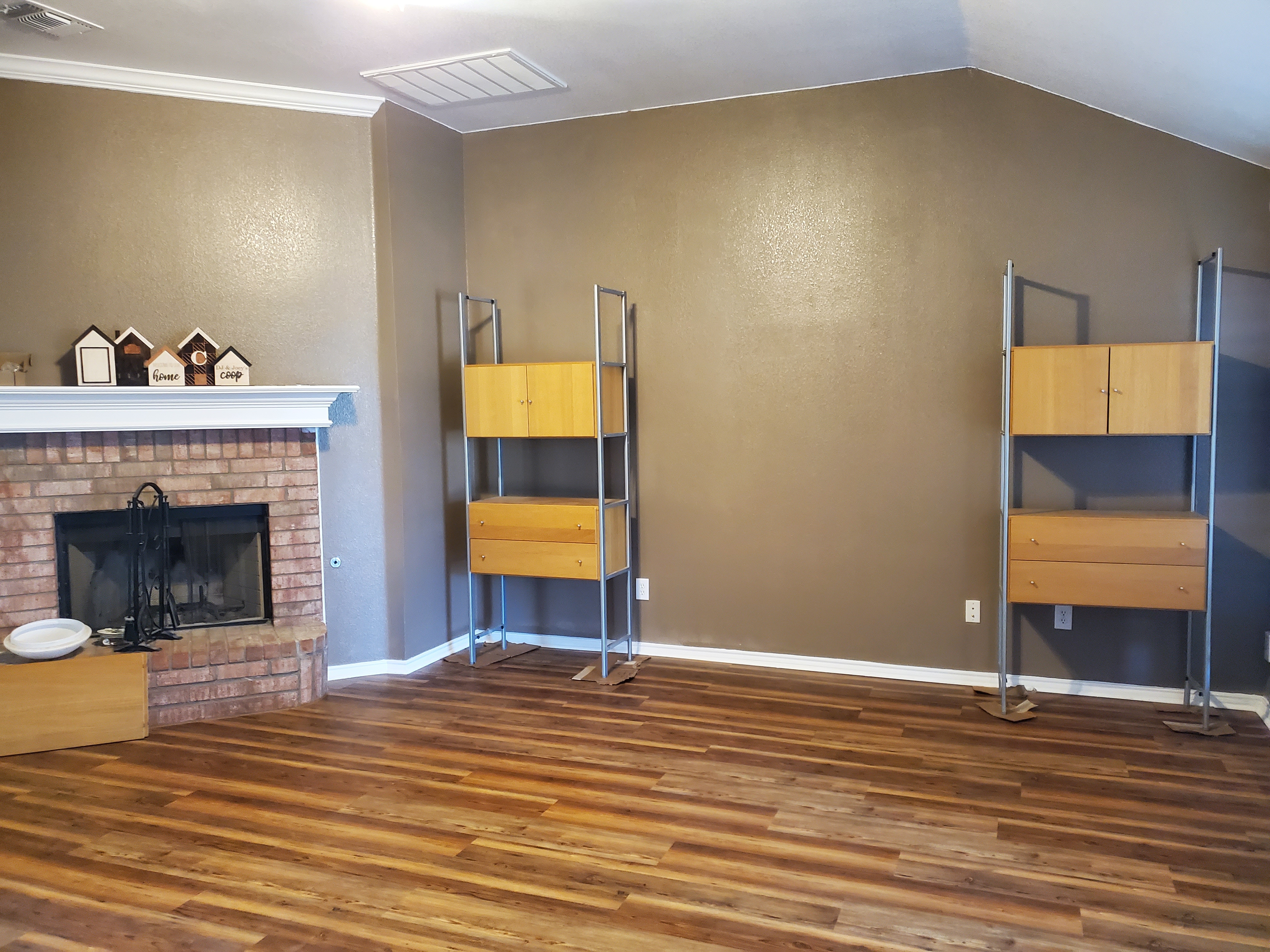 the image shows a living room with a fireplace and two modular shelving units. the fireplace is made of brick and has a white mantel with small decorative houses on top. the shelving units are made of wood and metal, and each one has a cabinet and drawers. the floor is made of wood planks. the walls are painted a neutral color.