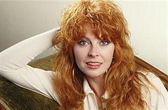 a photo of a woman with long, curly red hair. she is wearing a white shirt and is sitting and smiling for the camera. it appears to be a professional studio photo.