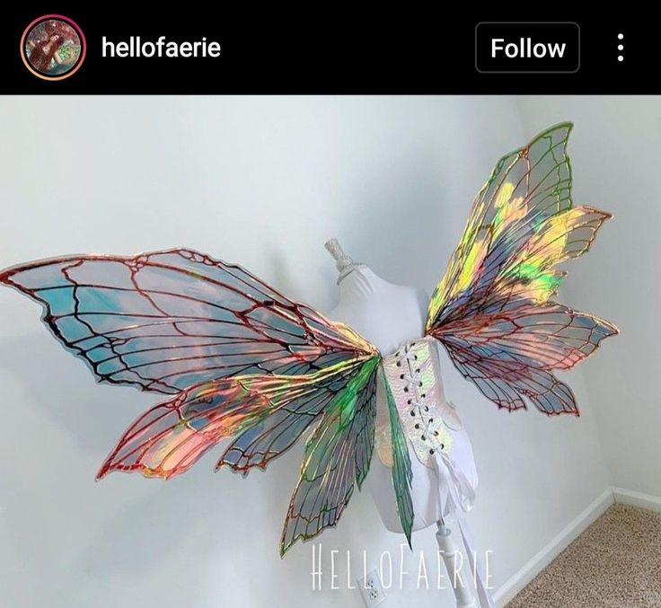 the image shows a pair of iridescent fairy wings attached to a white corset on a mannequin. the wings are intricately designed with iridescent colors and veined patterns. the overall aesthetic is ethereal and magical. the image appears to be a display or advertisement for a costume piece.