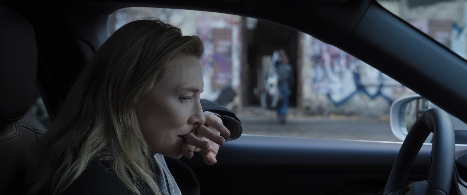 a woman with blonde hair is sitting in the driver's seat of a car, she looks contemplative with her fingers to her mouth as if deep in thought. the background suggests an urban environment.