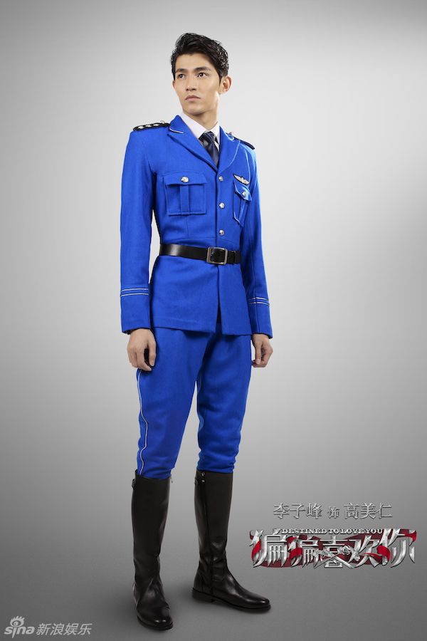 a man stands facing forward, wearing a bright blue militarystyle uniform and black boots. he has short black hair and is looking slightly to his left. the uniform consists of a jacket with pockets and a belt, and matching pants. the background is a gradient gray. there is text and logos at the bottom right and left corners of the image.