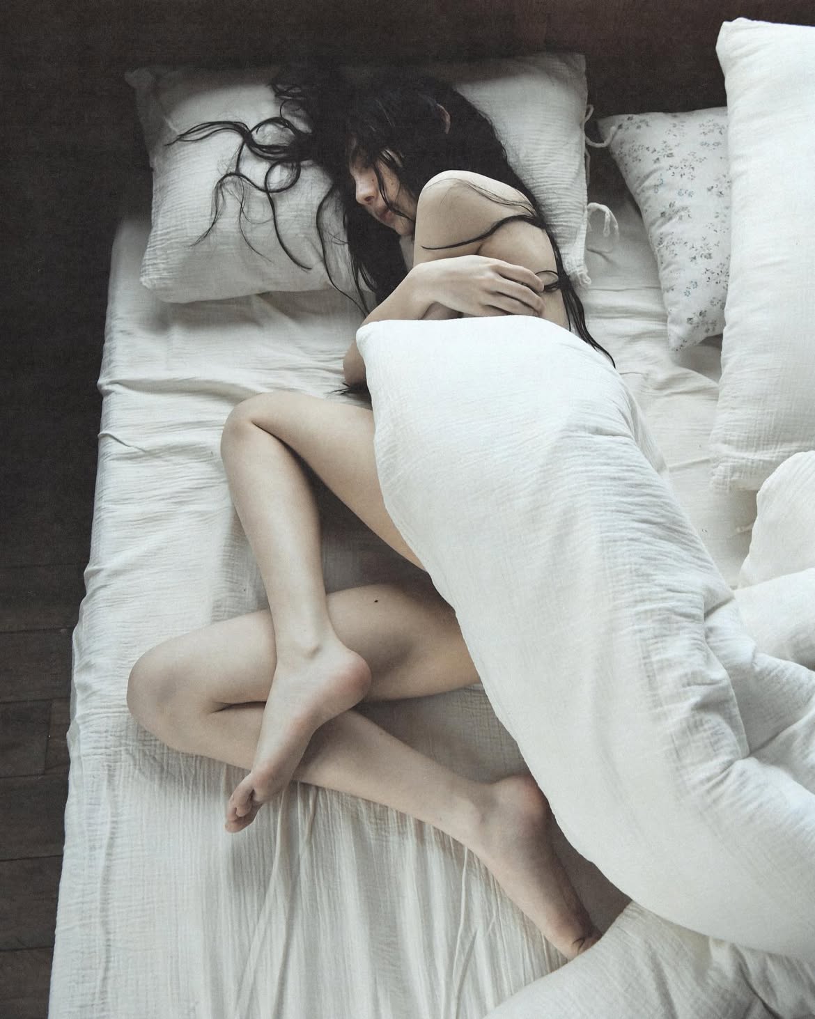 a woman with dark hair is curled up in a bed with white sheets. she is mostly covered by a white blanket and rests her head on a pillow. her legs are crossed, and the top view shows the entire scene within the bed.