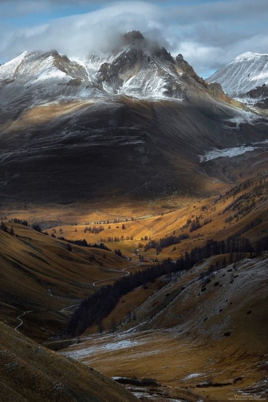 the image depicts a mountainous landscape with a deep valley running through it. the mountains are partially covered in snow, with clouds shrouding the peaks. a winding road snakes through the valley, which is filled with goldenhued vegetation and scattered trees. the sunlight creates dramatic shadows across the terrain, highlighting the ridges and folds of the landscape. the scene captures a sense of remote beauty and the changing seasons.