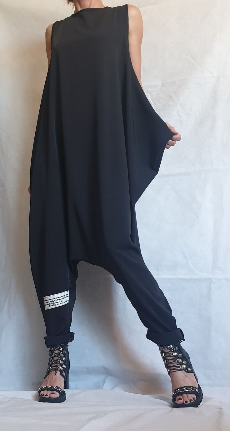 a woman is posing in a black sleeveless jumpsuit with a baggy fit, the jumpsuit has a belt around the waist, she is wearing black high heeled sandals with chain detail.