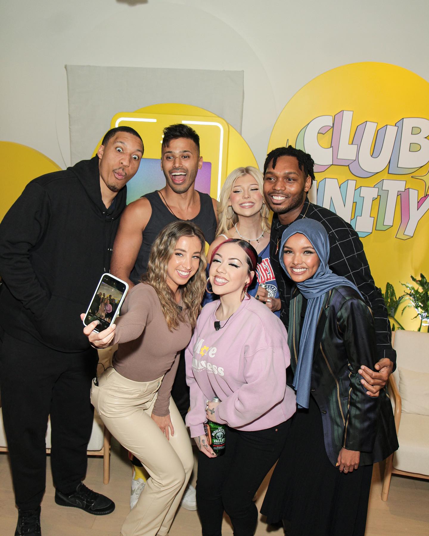 a group of seven people are standing together in front of a yellow backdrop that says "club nity" . they are smiling and posing for a photograph. one of the women is holding up a smartphone to take a selfie. there are both men and women in the group, and they are all dressed casually.