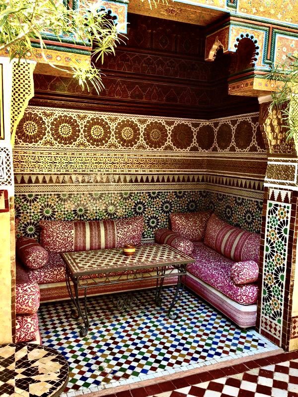 an ornate indoor seating area featuring traditional moroccan architecture. the walls are covered in intricate mosaic tilework with detailed ornamental patterns. a small table sits between two cushioned seating areas with patterned cushions. the floor has a checkerboard tile design in various vibrant colors.