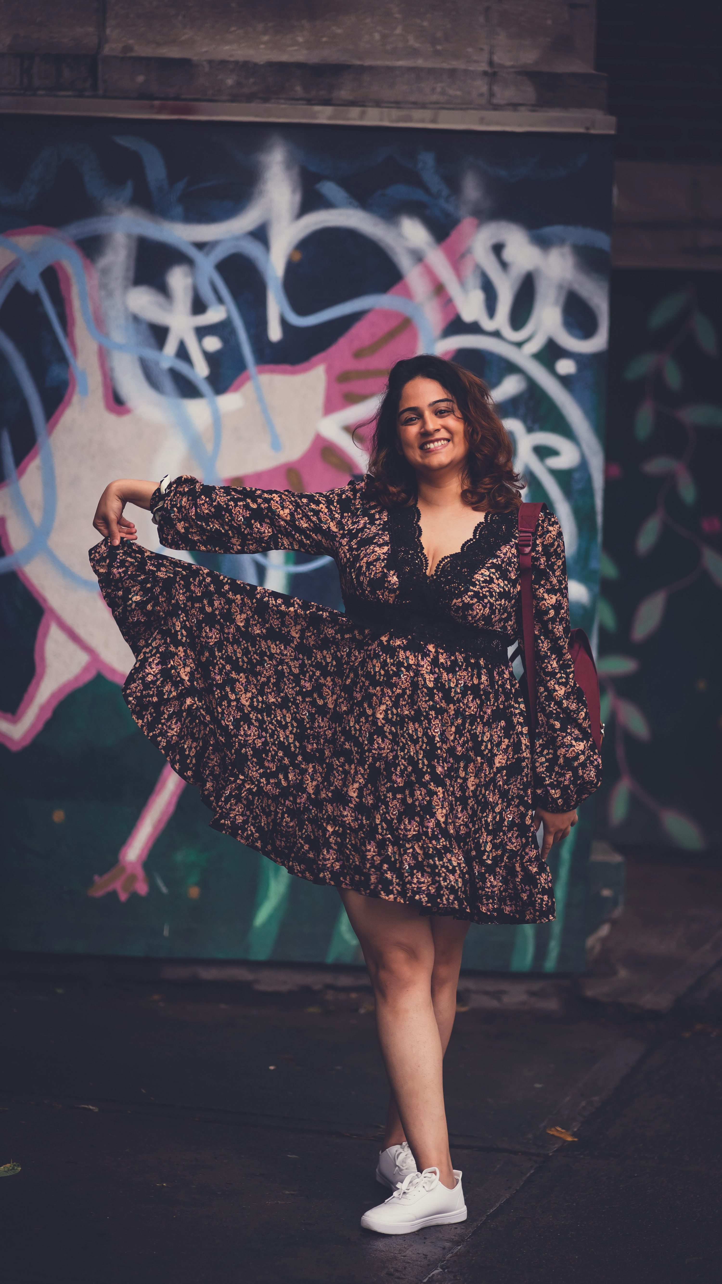 a young woman with long dark hair is standing in front of a graffiticovered wall. she's wearing a dark floral print short dress with long sleeves and a vneck, accented with lace. she has a burgundy shoulder bag slung across her body, and she's wearing white sneakers. she is smiling and her posture suggests confidence and playfulness. the overall style is urban and fashionable. the image is welllit and sharp.