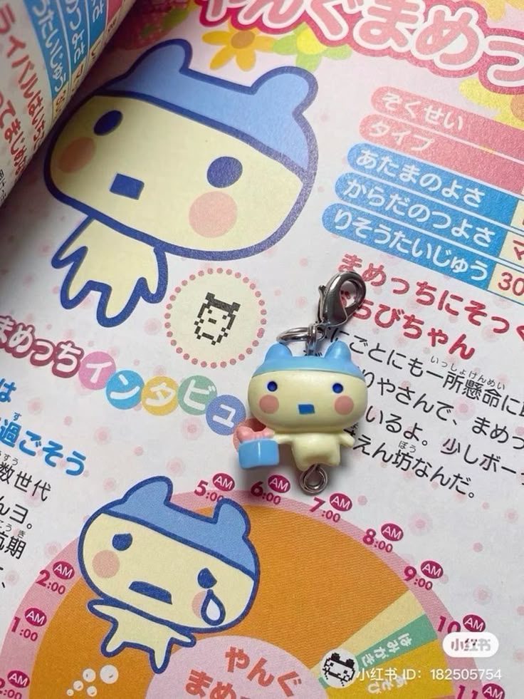 the image shows a page from a book featuring illustrations and text in japanese, with a tamagotchi charm placed on top. the illustrations depict a cartoon character with a blue hat, and there are also other design elements like a time chart and additional characters in smaller pixelated versions.