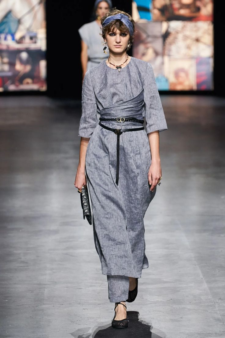 a fashion model walks the runway during a fashion show. she is wearing a long, grey dress with a black belt and shoes. she is accessorized with a blue headband, earrings, and a necklace. the model is walking in a casual yet elegant style.