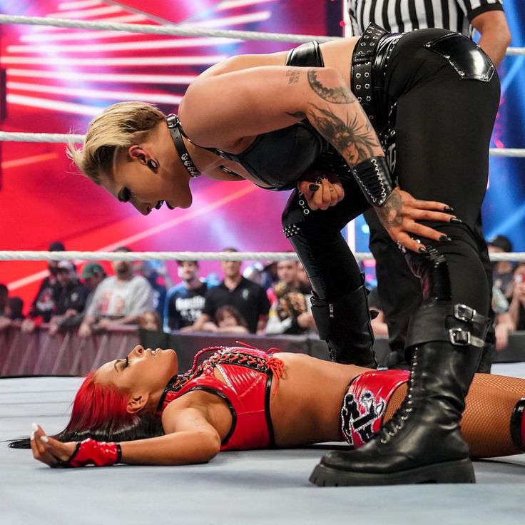 this image captures the intensity of a wrestling match between two female wrestlers. the wrestler in black leather attire is standing over the defeated wrestler in red, who is laying on the mat. both women are looking at each other, their expressions filled with a mix of determination and aggression. the wrestler in black is showcasing a powerful stance, highlighting her dominant position in the match. this image embodies the raw emotion and rivalry that defines professional wrestling.