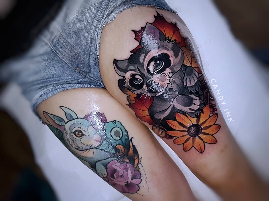 the image showcases two colorful tattoos on a woman's thighs.  the tattoo on the right thigh depicts a cute raccoon nestled amongst autumnal leaves and a sunflower. the tattoo on the left thigh features a whimsical rabbit with a blue feather and a pink rose. both tattoos are executed in a vibrant and detailed style.