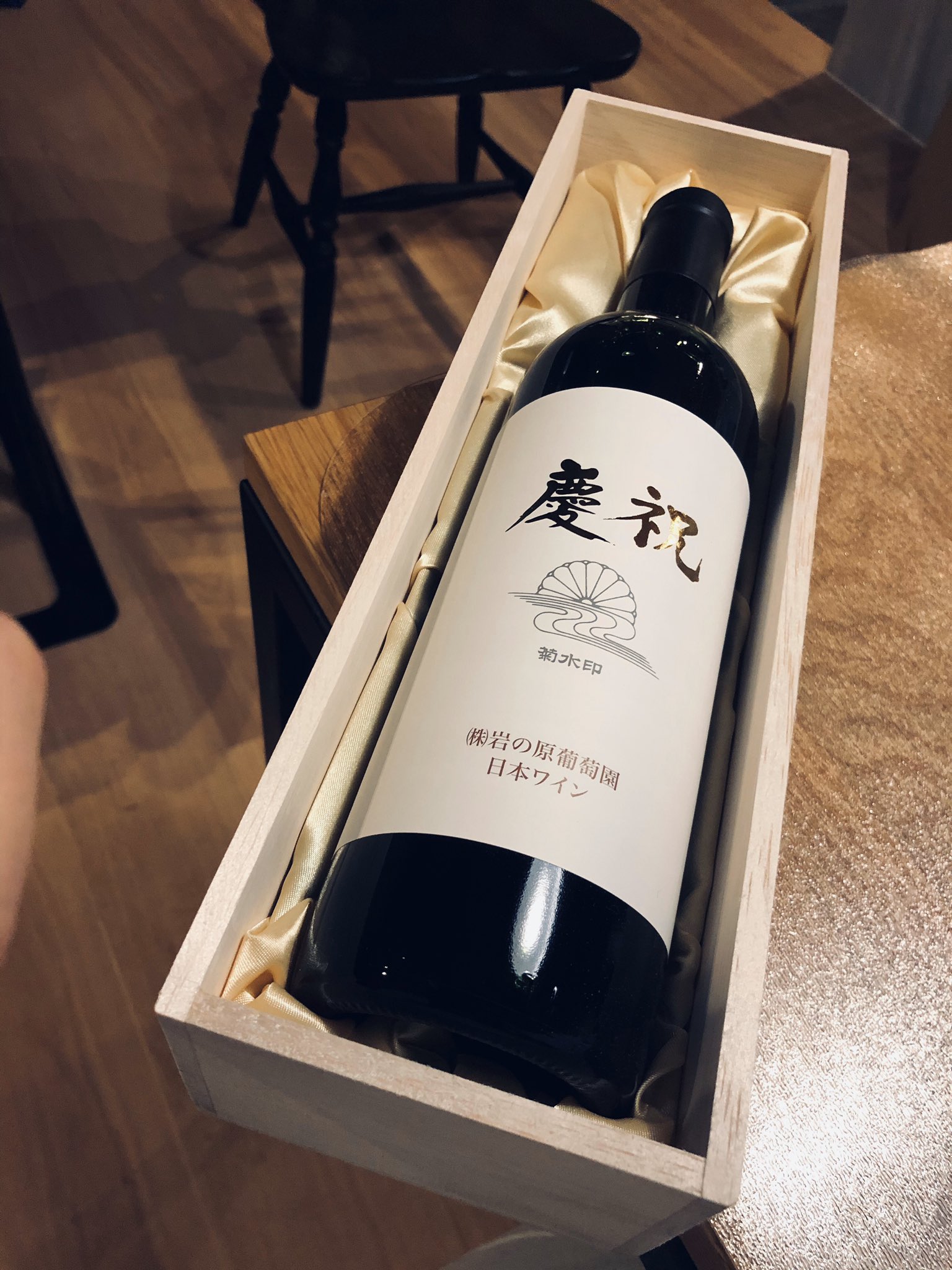 a bottle of japanese red wine in a wooden presentation box, resting on a hardwood floor. the box is lined with cream colored satin. the photo is taken from above.