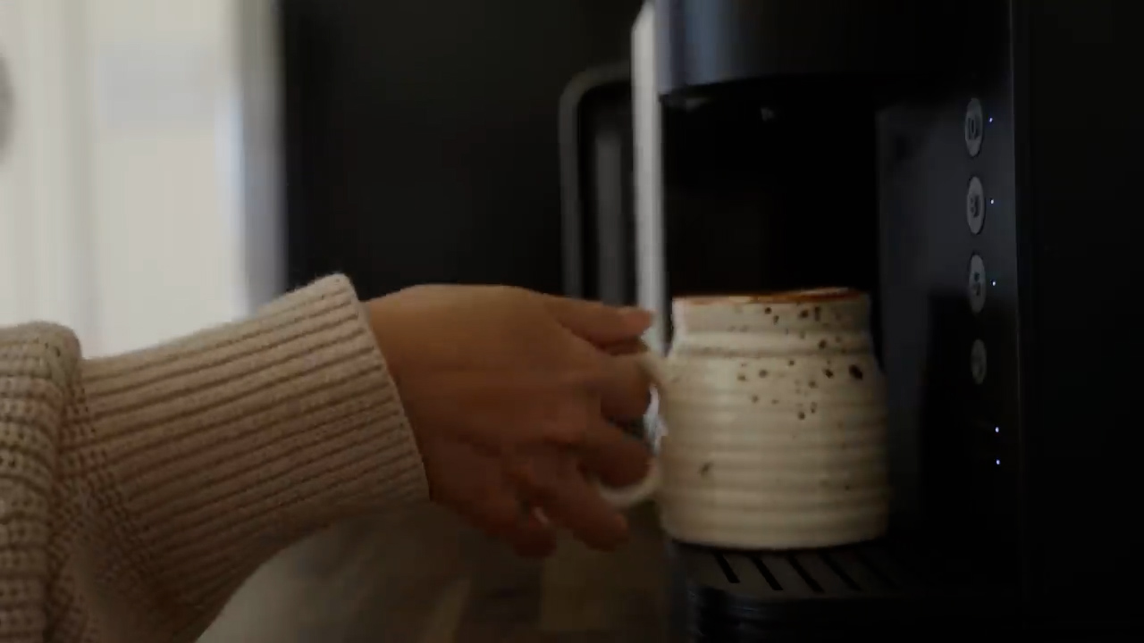 a person's hand, clad in a cozy, beige sweater, reaches out to grasp a speckled, creamcolored mug that is positioned beneath the spout of a sleek, black coffee maker. the mug is filled with a steaming cup of coffee, hinting at a cozy and inviting morning routine. the background features a neutral color palette with soft lighting.