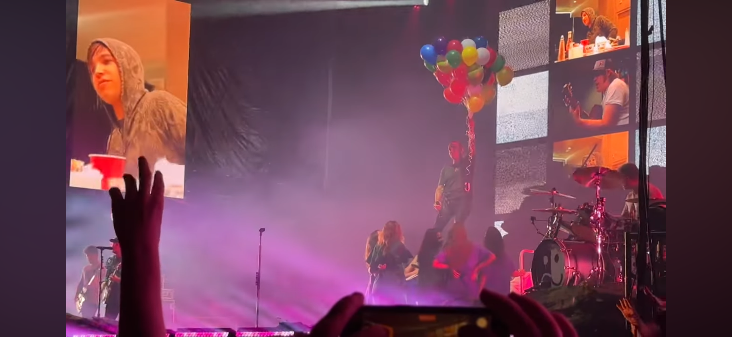 this is a live concert with a band playing on stage. the band includes a drummer, guitar player, and singer. the singer is holding a bunch of balloons in the air.  the audience is visible in the foreground, cheering and enjoying the performance.  the stage is lit up with vibrant lights, and the concert is happening in an indoor venue.  the concert appears to be taking place at night.  the energy of the show is electric, and the band is giving a passionate performance.