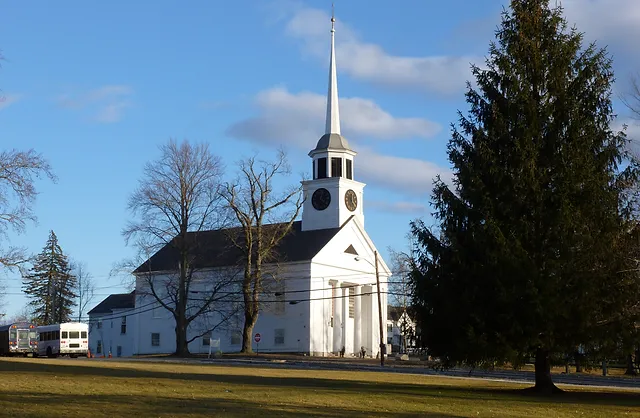 a full shot shows a white church with a steeple surrounded by green grass and trees under a cloudy blue sky. the church has a clock tower with a clock face on each side. to the left of the church is a white and red bus parked on the street. the lawn is yellowgreen and there are several bare trees near the church. a large evergreen tree stands to the right of the church.