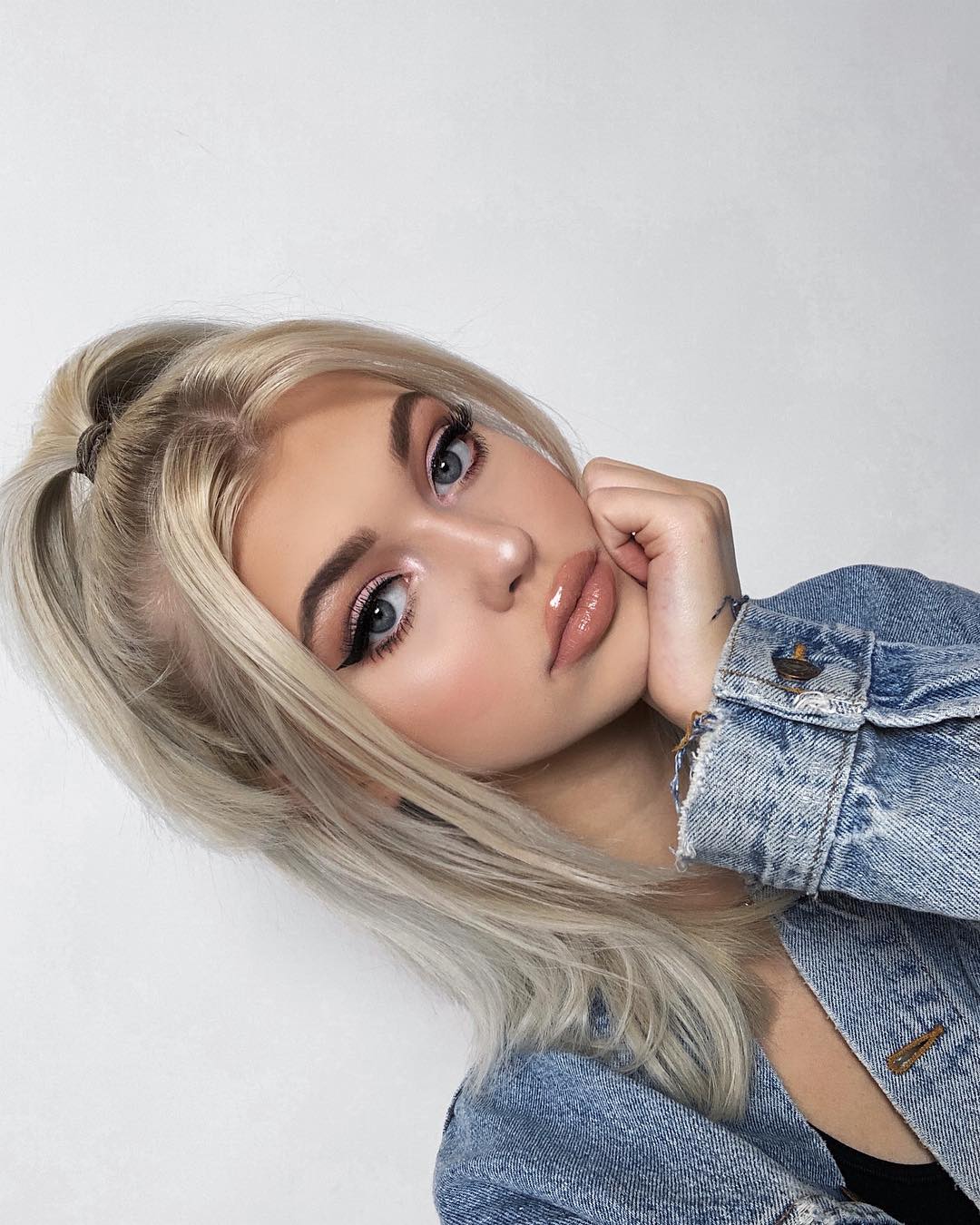 a young woman with blonde hair, light skin, and blue eyes is looking at the camera. she is wearing a denim jacket and has makeup on, including eyeliner and lipstick. her hair is styled in a ponytail. she is posing with her hand near her face. the background is white.