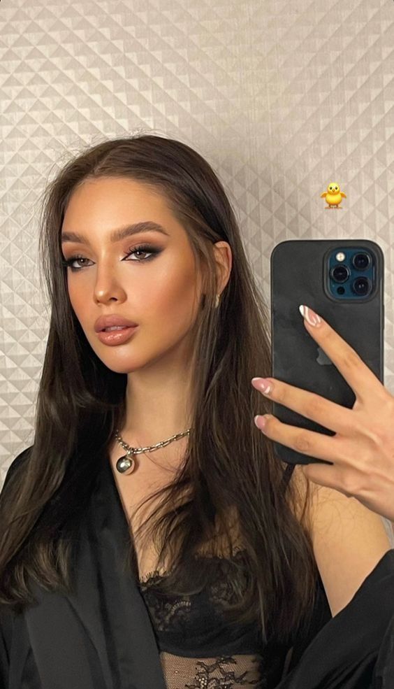 a beautiful young woman with long dark hair is taking a selfie with her phone. she is wearing a black top with a silver necklace and has dark makeup on her eyes. she is smiling slightly and looking directly at the camera. the image is taken indoors with a plain background.