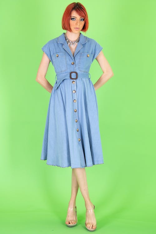 a woman stands in front of a green background wearing a light blue dress with a brown belt, a necklace, and clear heels. she has red hair.
