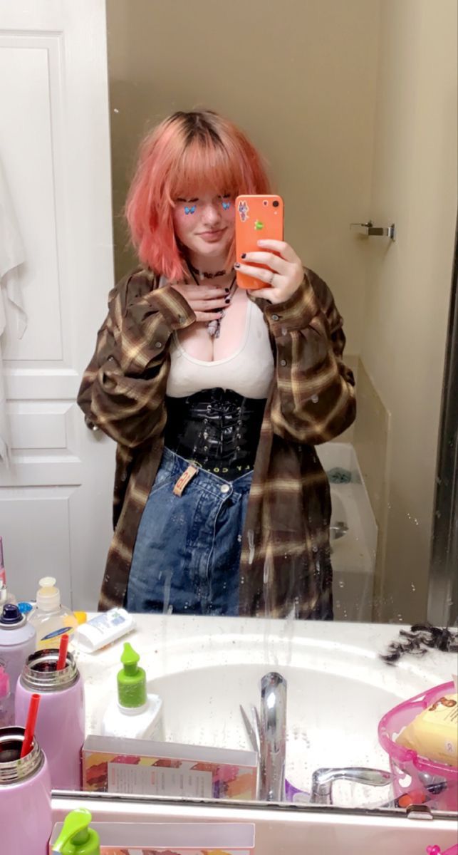 a woman with pink hair and decorative blue eye makeup takes a selfie in a bathroom mirror. she wears a brown and tan plaid shirt over a white top, a black corset, and a denim skirt. she has several necklaces. the bathroom sink is cluttered with various items including water bottles.