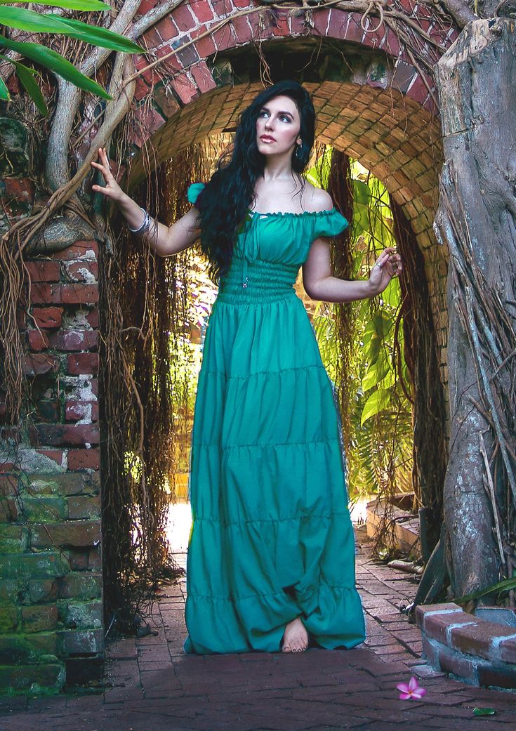 a fulllength portrait of a young woman with long dark hair wearing a flowing teal maxi dress stands under a brick archway overgrown with vines and tropical plants.  she is barefoot and appears calm and serene. the setting is lush and somewhat mystical.  the image has a dreamy, fairytale quality.