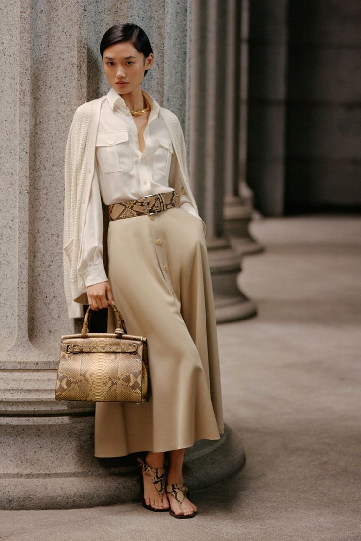 a fullbody shot of a fashion model standing elegantly between two columns, wearing a stylish beige outfit. the outfit includes a silk shirt, a beige cardigan, a highwaisted midi skirt, and a snakeskin belt. she's carrying a snakeskin handbag and wearing snakeskin sandals. the model is wearing gold jewelry, including a necklace. the overall style is sophisticated and chic. the background is an urban setting, with columns creating an architectural backdrop. the color palette is neutral and pastel.