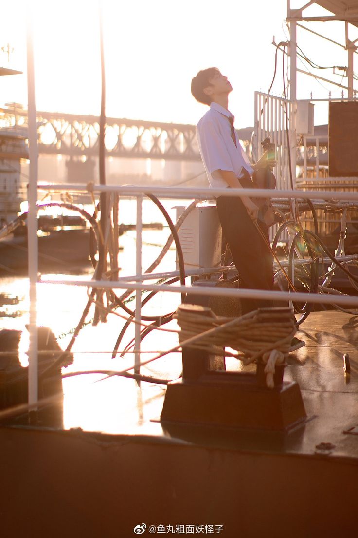 a young man, dressed in a white shirt and dark pants, stands on a platform by a river, looking up towards the setting sun. the warm, golden light of the sunset illuminates the scene, creating a serene and peaceful atmosphere. a bridge is visible in the background, and the water of the river reflects the colors of the sky. the overall impression is one of tranquility and contemplation.