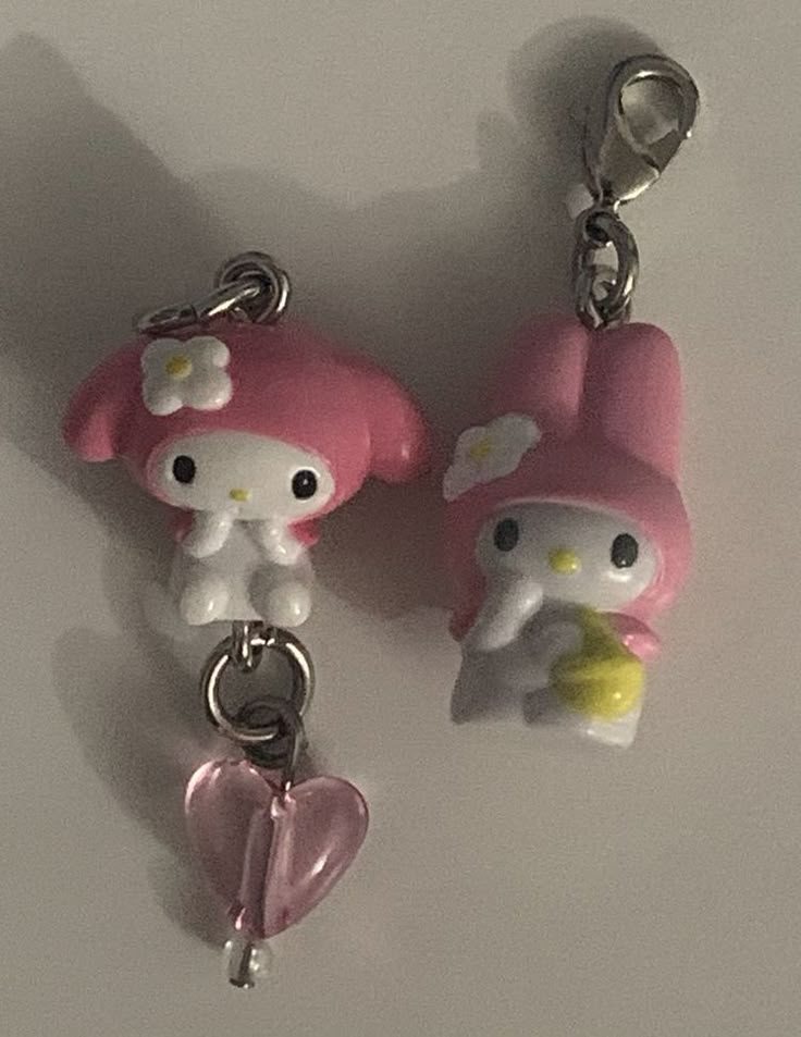 two my melody character keychains/charms are displayed. one features my melody holding a yellow object, attached to a silver lobster clasp. the other features a my melody figure connected to a pink heart charm by silver jump rings. the background is a plain, light surface.