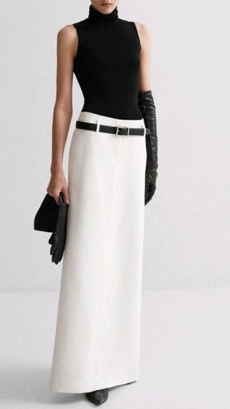 a fashion model poses in a white skirt, black turtleneck, and black gloves. she has a simple belt and the outfit is very elegant and stylish. the minimalist design of the outfit makes it easy to wear and flattering for almost any figure. the background is a plain white wall which helps to draw attention to the outfit.
