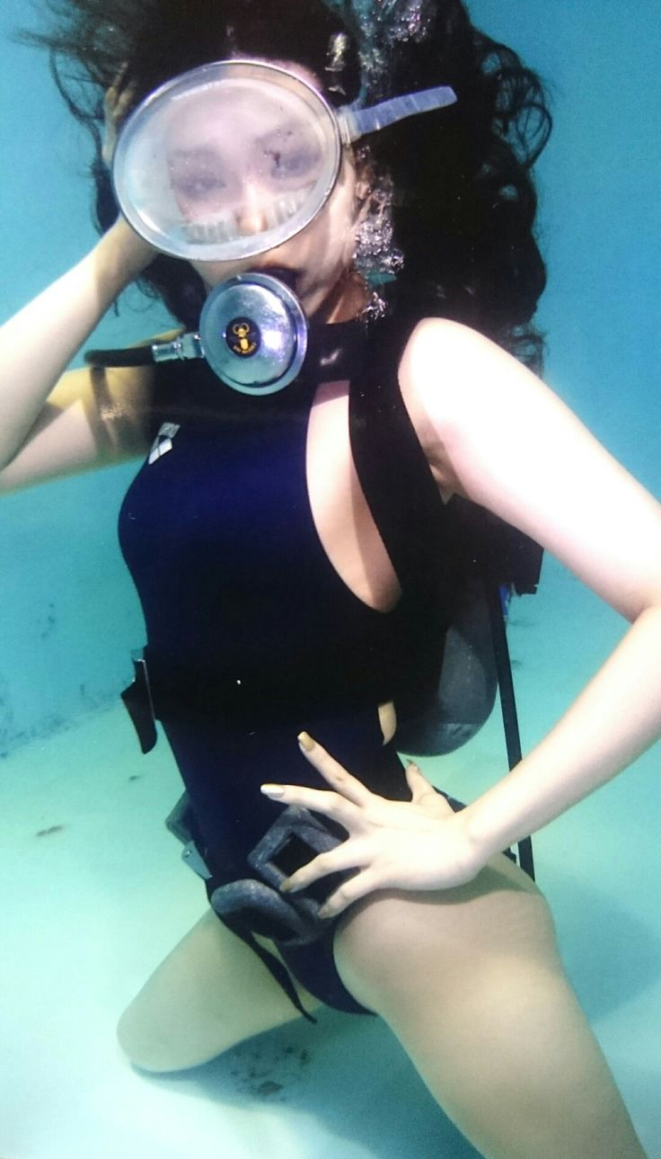 a woman wearing a black swimsuit, scuba gear and a mask, is underwater with her body partly out of the water, she is looking at the camera. her arms are outstretched and she is posing.