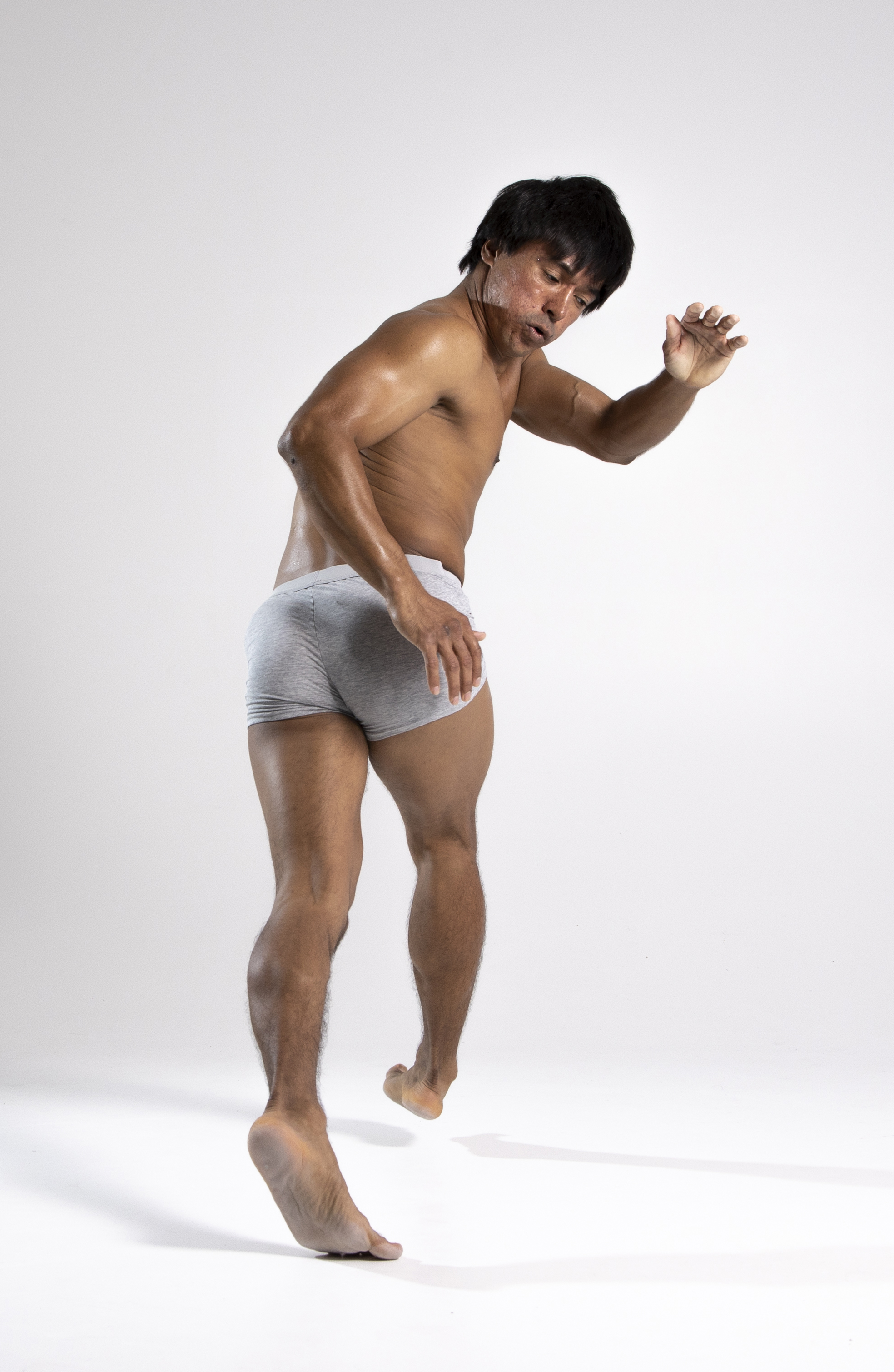a barechested asian man wearing gray shorts is seen from a back view, barefoot and walking. his right hand is extended slightly forward as if reaching out. the background is a bright, plain white.