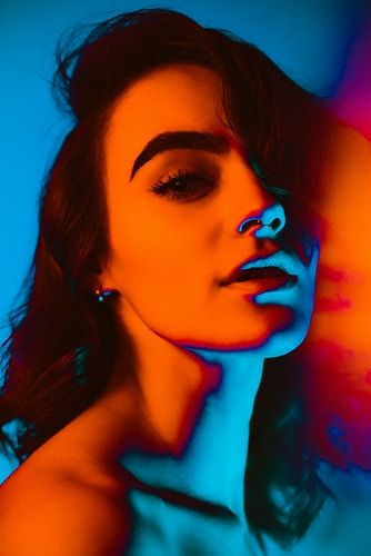 a portrait of a woman with strong color lighting. the lighting is predominantly orange on the face and shoulder with blue outlining the neck and parts of the face, creating a dramatic effect. her hair is dark, and she is looking slightly up and to the side.