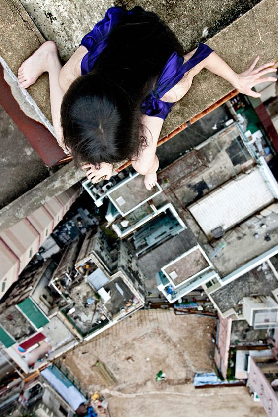 a woman in a blue dress sits on the edge of a building, high above the city. she looks down with a mixture of fear and awe as she contemplates the height and the danger of her situation. the perspective of the photograph is from her point of view, looking down at the city below. the image captures the feeling of vertigo and the thrill of facing one's fear.