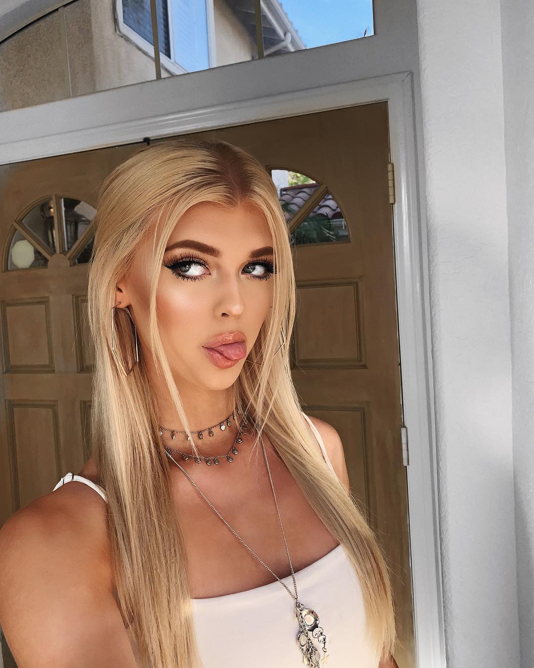 a woman with blonde hair is posing for a selfie with her tongue out. she is wearing a white top, a necklace, and earrings. she is standing in front of a doorway.