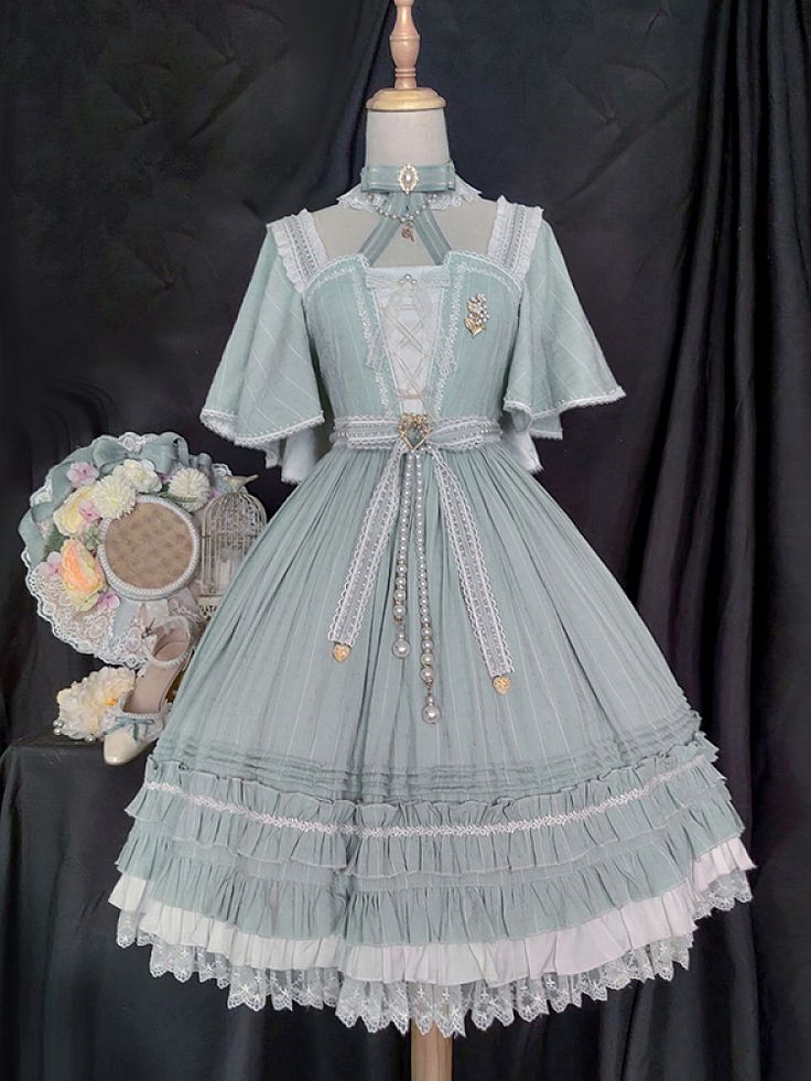 a fulllength image of a mint green lolita fashion dress displayed on a mannequin. the dress is made of a light, flowy fabric and features intricate lace detailing, a layered skirt with ruffles, and delicate pearl embellishments. the bodice is fitted with a sweetheart neckline and short puffed sleeves. a matching hat with flowers rests on a stand behind the dress, further emphasizing the vintageinspired aesthetic.