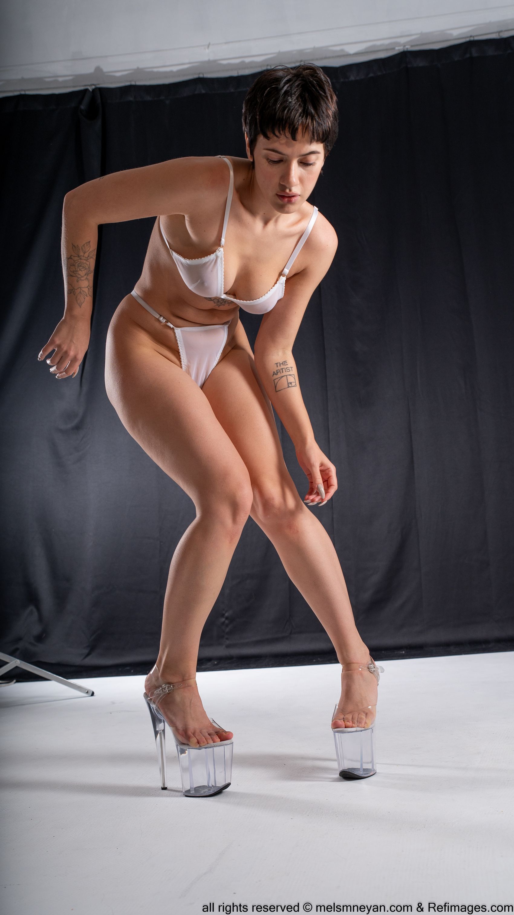a fullbody studio shot depicts a woman wearing white lingerie and clear platform heels. she has short hair and a tattoo on her left arm, and is looking down. the background is dark.