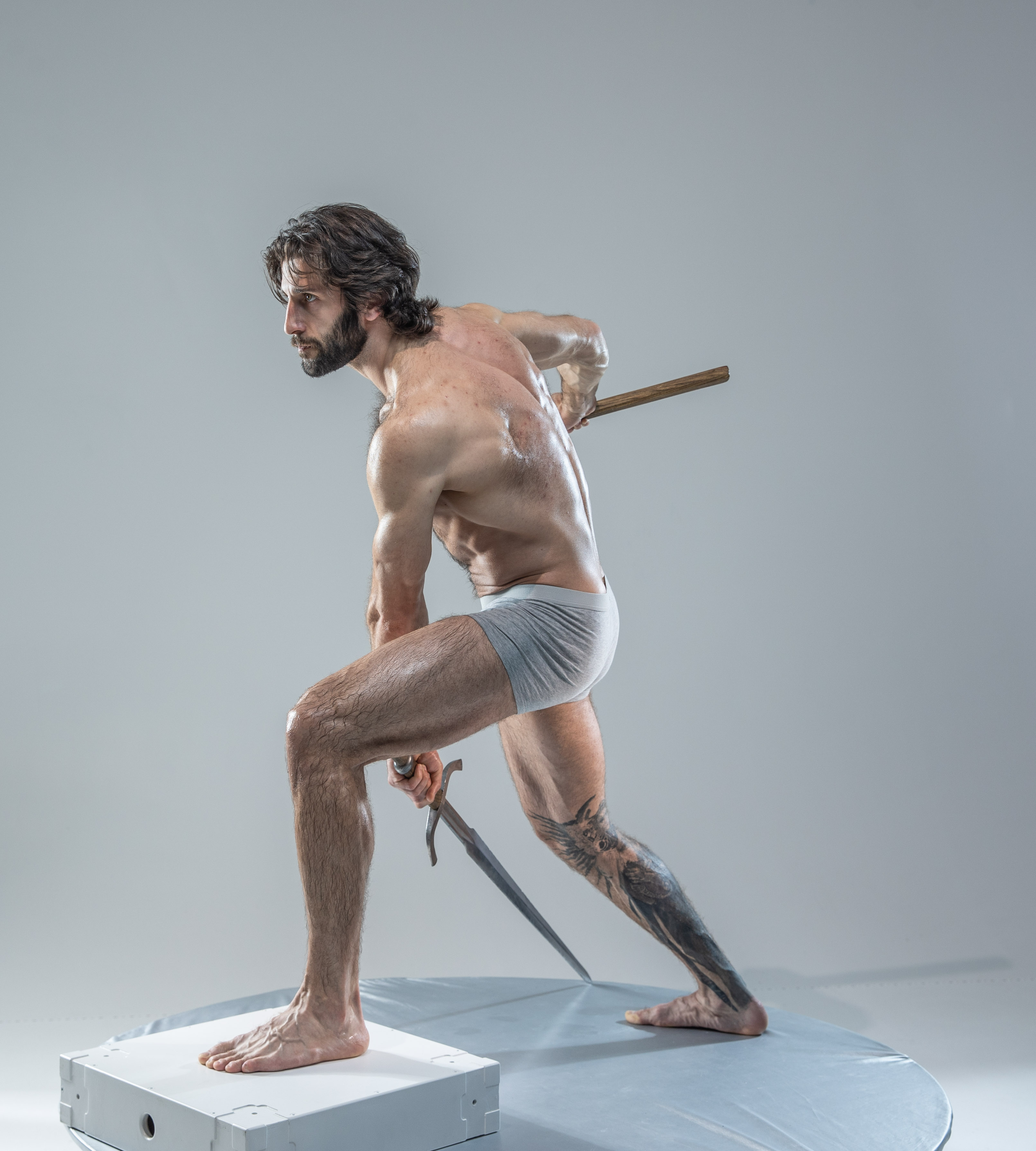 a muscular man with long hair is dynamically posed on a small platform in a photography studio.  he is wearing short underwear, holding a sword in his right hand, and a shorter weapon or staff in his left hand, both pointing behind him.  he is barefoot and has tattoos on his left leg.  he appears to be engaged in combat.  the background is solid gray and the floor is a light bluegray.