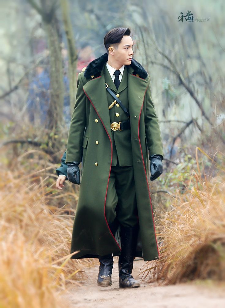 a man dressed in a long green military uniform with red trimming, black gloves, and black boots is walking on a dirt path in an outdoor environment. the background consists of foliage, grass, and trees, suggesting a forest or similar natural setting. the man has a dark tie and a black fur collar on his coat. the coat is buttoned, and he is wearing a belt with a distinctive buckle.