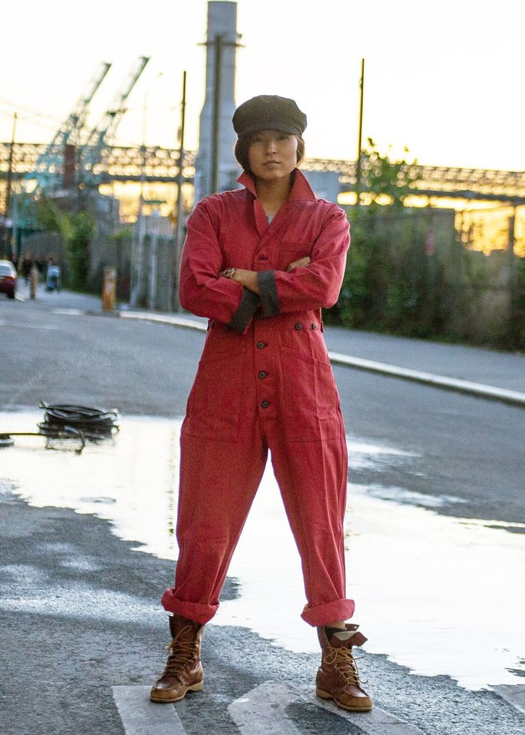 a full shot of a woman wearing a red jumpsuit, brown boots, and a newsboy cap stands confidently in an urban industrial setting.  the background features cranes, a dock, and industrial buildings at sunset.  she has her arms crossed and a serious expression on her face.  the overall mood is stylish and strong.