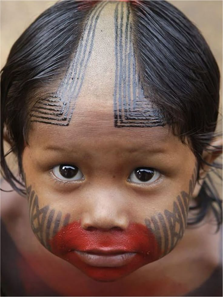 a closeup portrait of a young child with distinctive tribal markings and face paint. the child has dark hair and expressive eyes. there are black lines and geometric patterns painted on the forehead and cheeks, with a swath of red paint covering the mouth and lower part of the face. the hair is partially shaved on top, revealing a lightcolored patch with further black line markings.