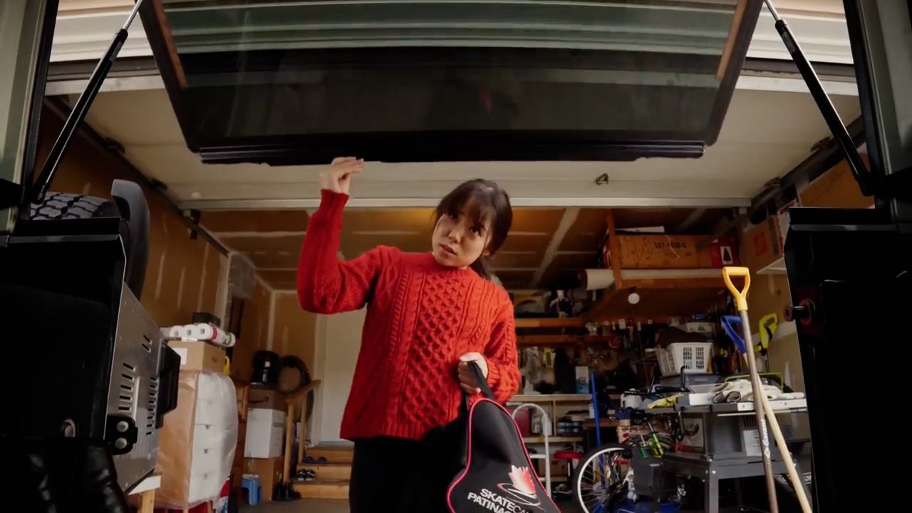 a young woman in a red sweater stands in a cluttered garage, reaching up to close the trunk of a car. the car trunk is open and takes up the top portion of the image, with the camera positioned low and looking up towards it. the woman holds a black and red sports bag slung over her shoulder. the garage is filled with various items, including tools, boxes, and sports equipment. the lighting is bright, likely from artificial sources.