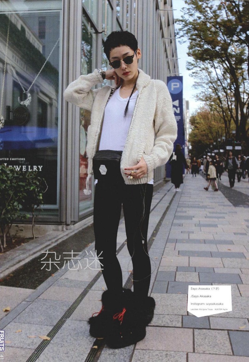 a young woman with short hair, sunglasses, and a black and white outfit, stands on a city sidewalk posing for a street style photo. she is wearing a creamcolored oversized cardigan, a white tshirt, black leggings, and black platform shoes. she has a black fanny pack slung across her chest and is also wearing a silver chain.  the background features storefronts, trees, and other pedestrians.  the image has a slightly desaturated, cooltoned color palette, reflecting the overcast daylight.