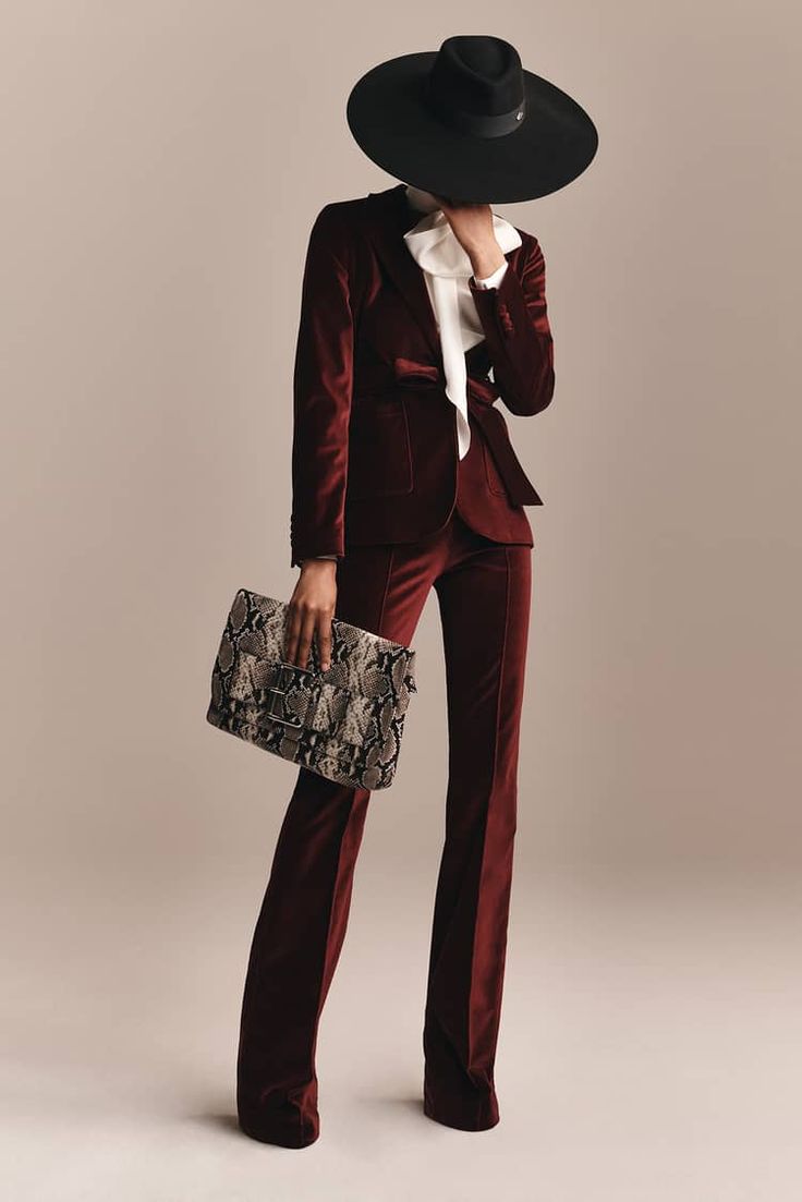 a woman is posing in a studio setting. she is wearing a black hat, a red velvet blazer, matching red velvet trousers, and a snakeprint clutch. the woman has an elegant and chic style.
