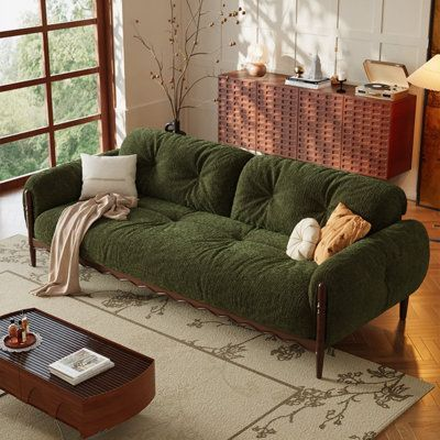 this image features a living room with a green tufted couch adorned with pillows and a draped blanket. a wooden coffee table with objects sits in front of the couch on a patterned rug. in the background, a wooden cabinet with decor items including a record player and vases is visible, illuminated by a lamp. a large window allows natural light to fill the room, enhancing the interior design and creating a cozy atmosphere.