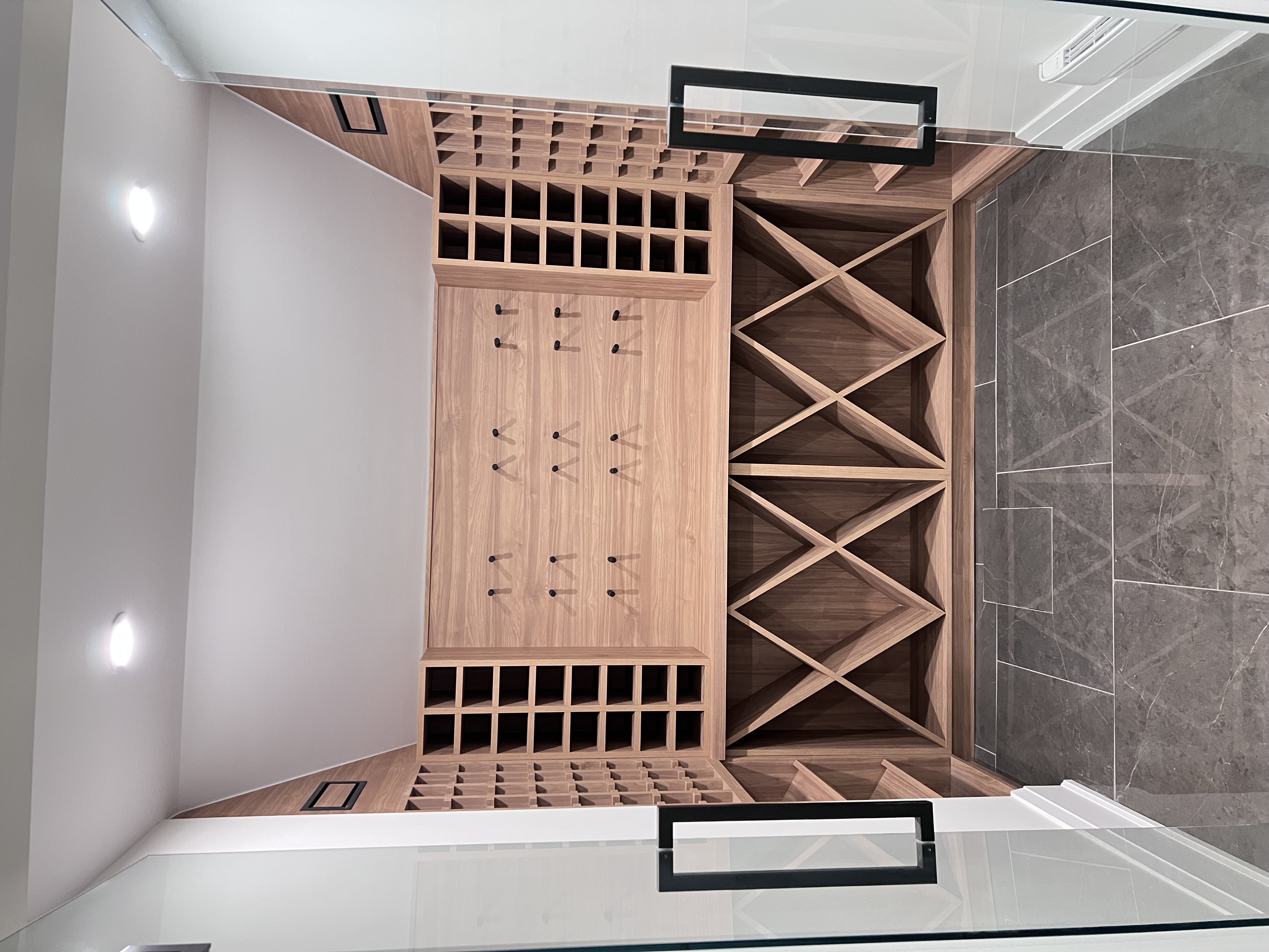 his image shows a modern, custombuilt wine cellar nestled in an attic space with a sloped ceiling. the wine storage features a combination of traditional and modern design elements.  the wine racks are primarily made of lightcolored wood, with a combination of cubbystyle storage for bottles and more open, xshaped racks. the floor is finished with sleek, large grey tiles.  recessed lighting is installed in the ceiling. glass doors are visible on either side of the cellar, suggesting access from another part of the house.  the overall aesthetic is clean, sophisticated, and luxurious.