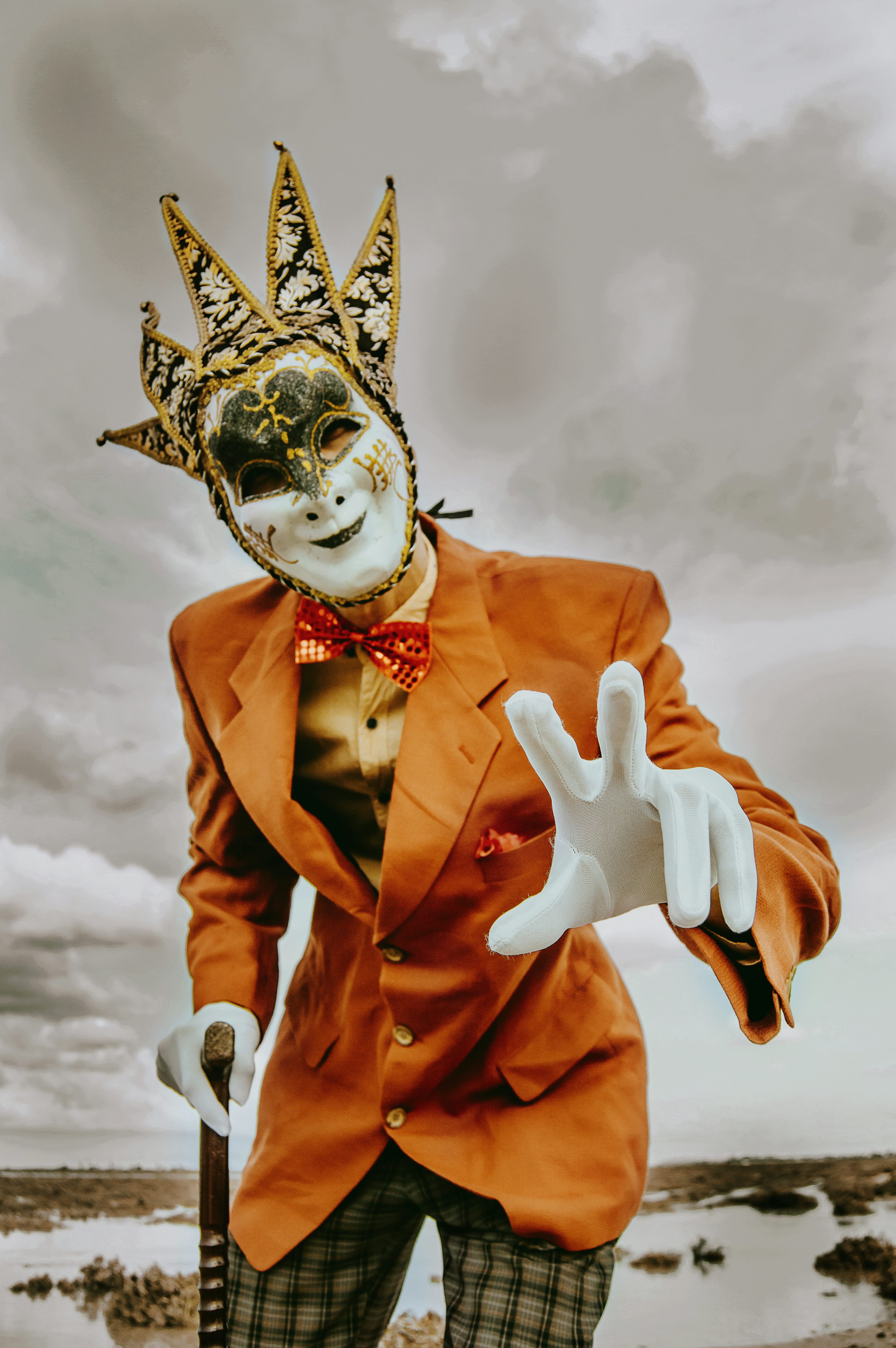 the image shows a man wearing a striking ornate venetian jester mask with gold and black detailing. he is dressed in a vintagestyle orange suit with plaid pants. he holds a walking cane in one hand and extends the other hand towards the viewer in a gesture that seems both inviting and slightly ominous. the background is a moody, overcast sky and desolate, watery landscape. the overall mood is dramatic, theatrical, and slightly mysterious, blending elements of whimsy and a sense of character. the lighting enhances the contrast and highlights the costume details.