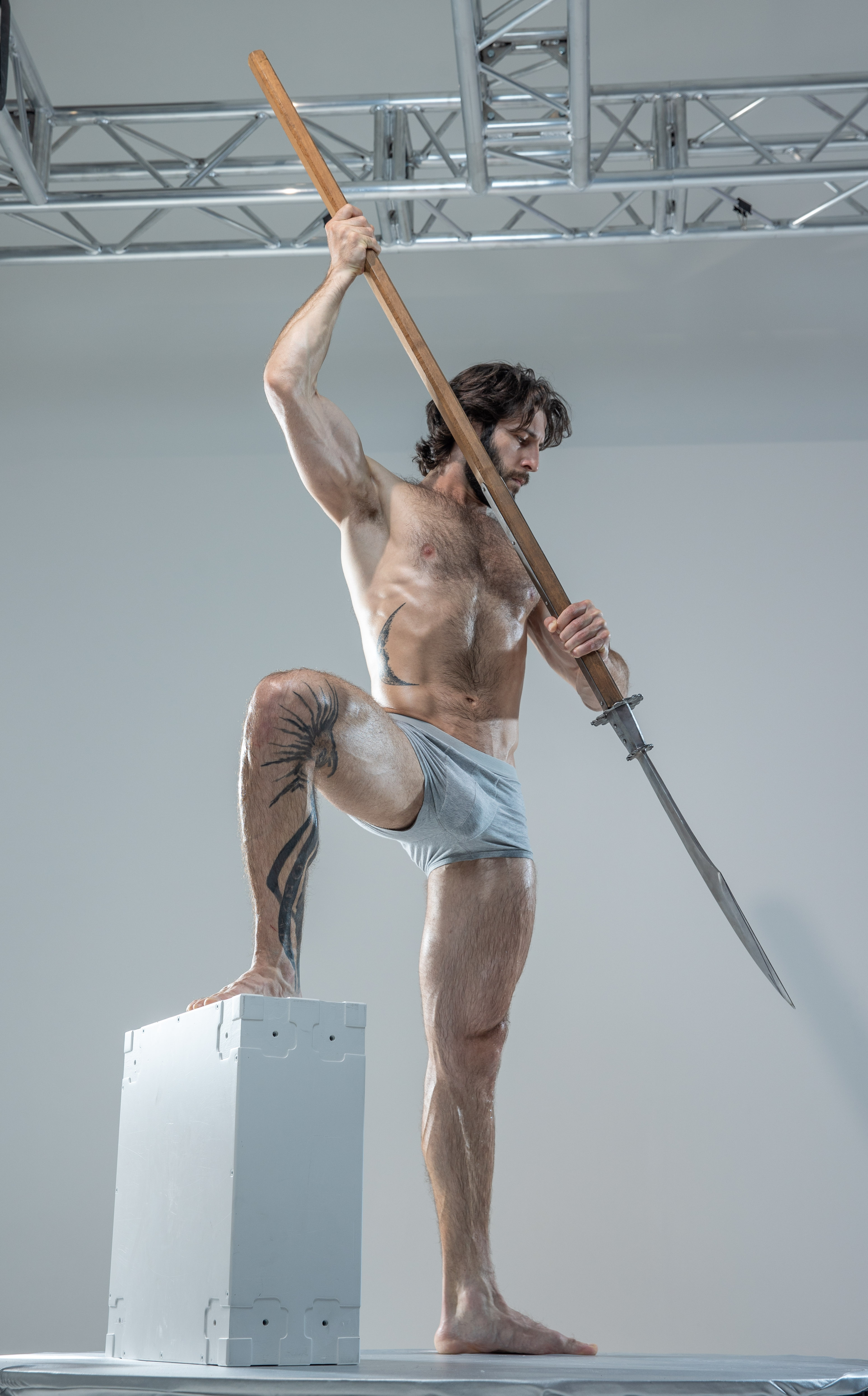 a muscular man with tattoos is posing on a white cube in a studio setting. he is holding a spear and is covered in white powder. he is shirtless and wearing grey underwear. the background is white and there is a metal structure visible. the lighting is artificial and the perspective is from a low angle.