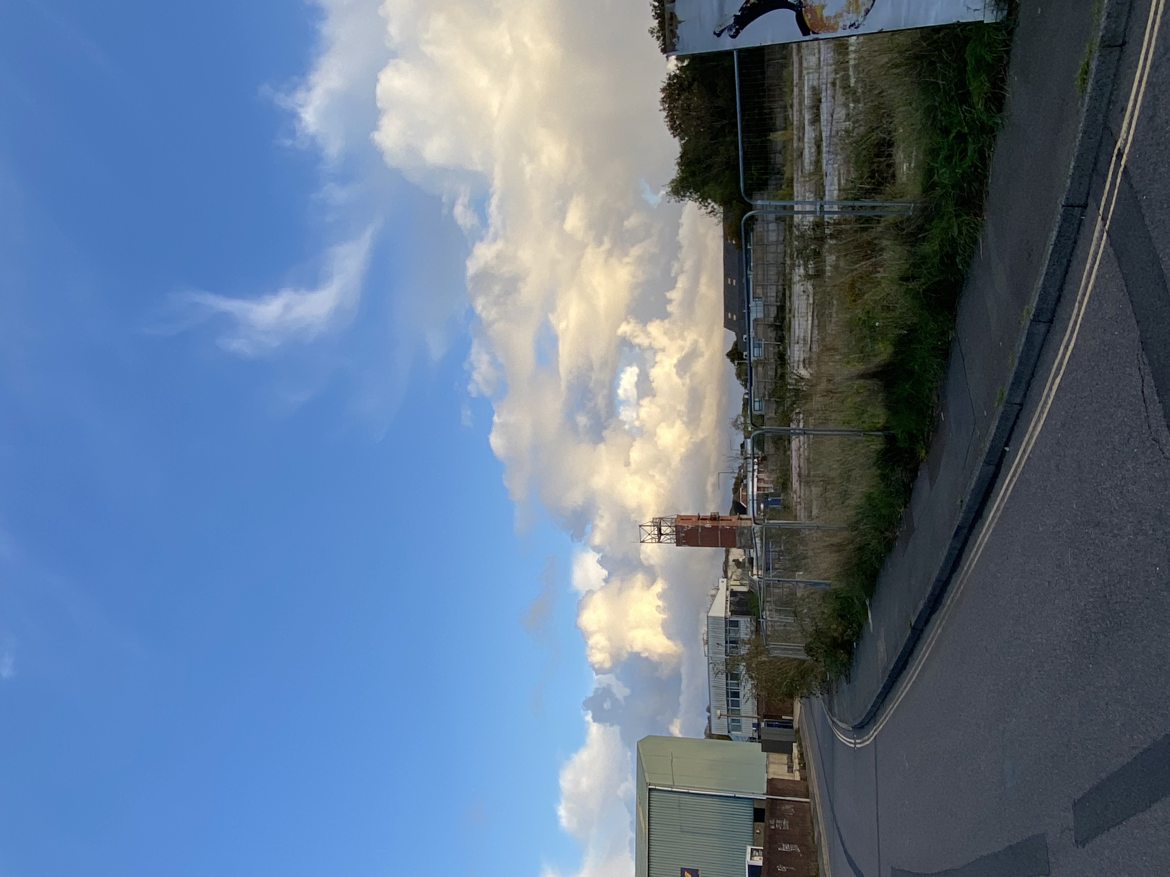 an empty, abandoned lot with a brick tower in the background. the road curves to the left with a sidewalk and grassy areas. the sky is a clear blue with fluffy white clouds.