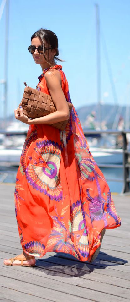 a young woman in a bright orange and floral maxi dress walks on a wooden deck overlooking a waterfront. she is smiling and has on brown sandals and sunglasses. she is carrying a brown handbag.  the woman is enjoying a relaxing day outdoors on vacation.