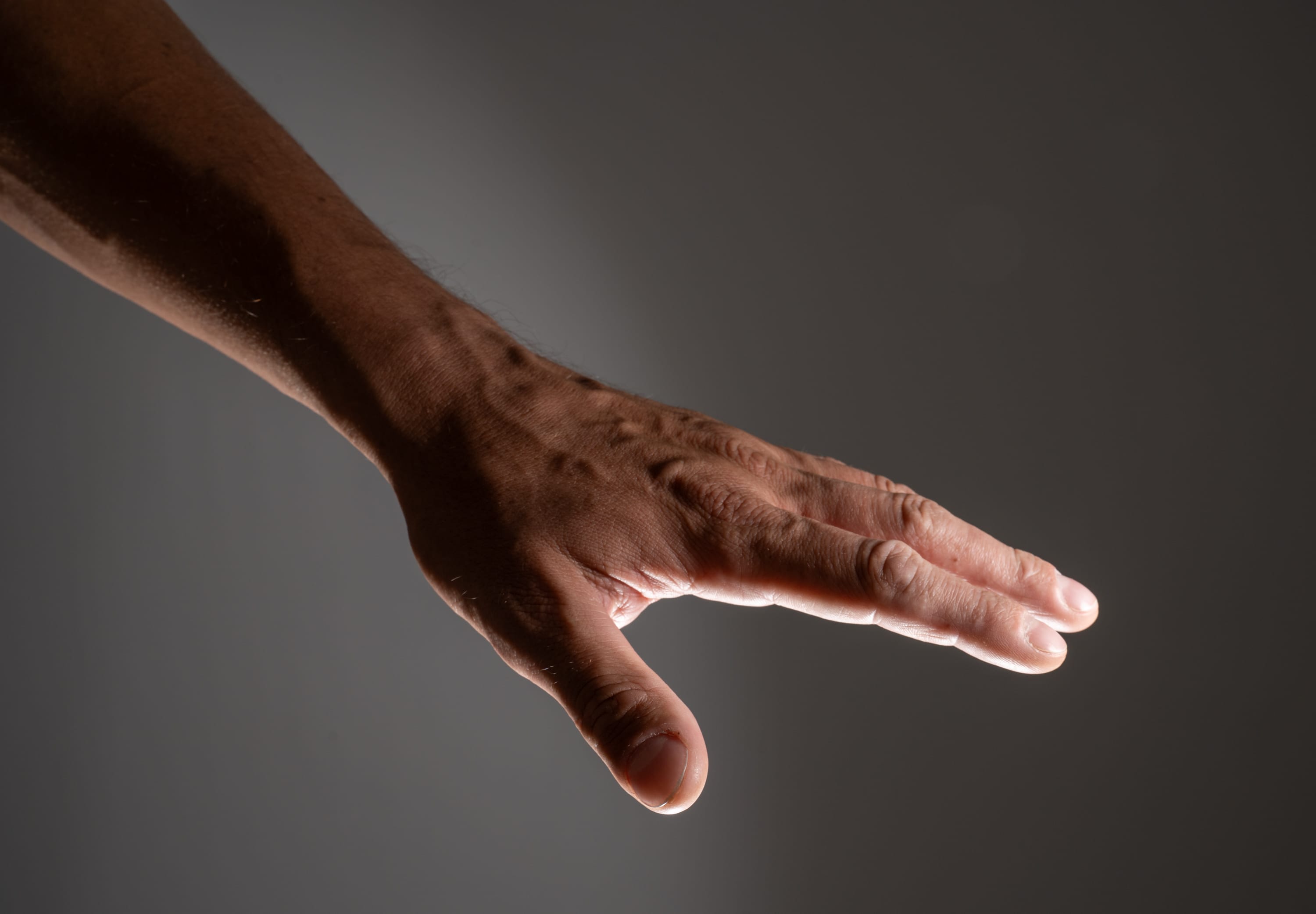 a closeup side view of a man's left hand and arm reaching forward. the hand is outstretched with the fingers slightly spread apart, as if reaching for something. the arm is muscular and the skin is tanned with visible veins. the lighting is soft and creates shadows on the hand and arm, highlighting the contours. the background is a dark gray color.