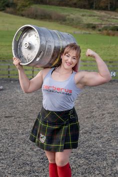a woman dressed in a kilt and tank top smiles confidently as she lifts a large barrel over her head.